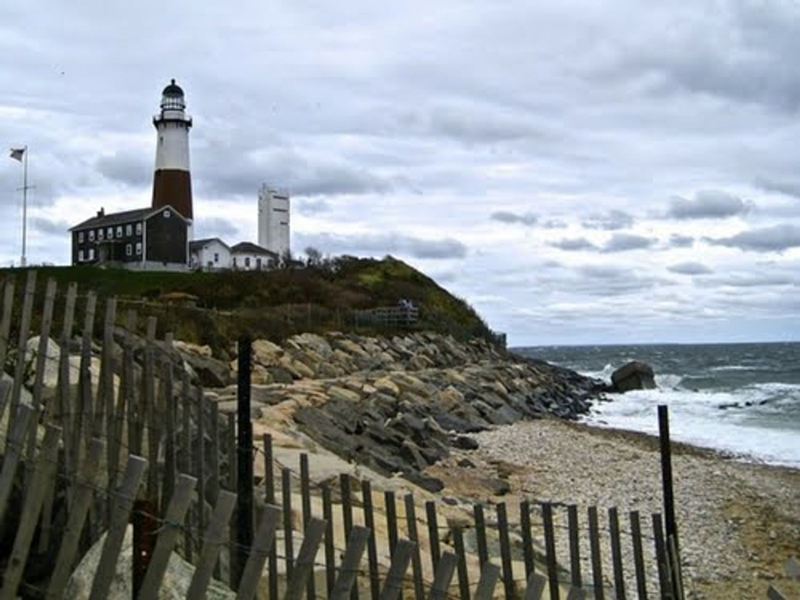 Lighthouse on rocky coastal cliff with wooden fence and beach below.