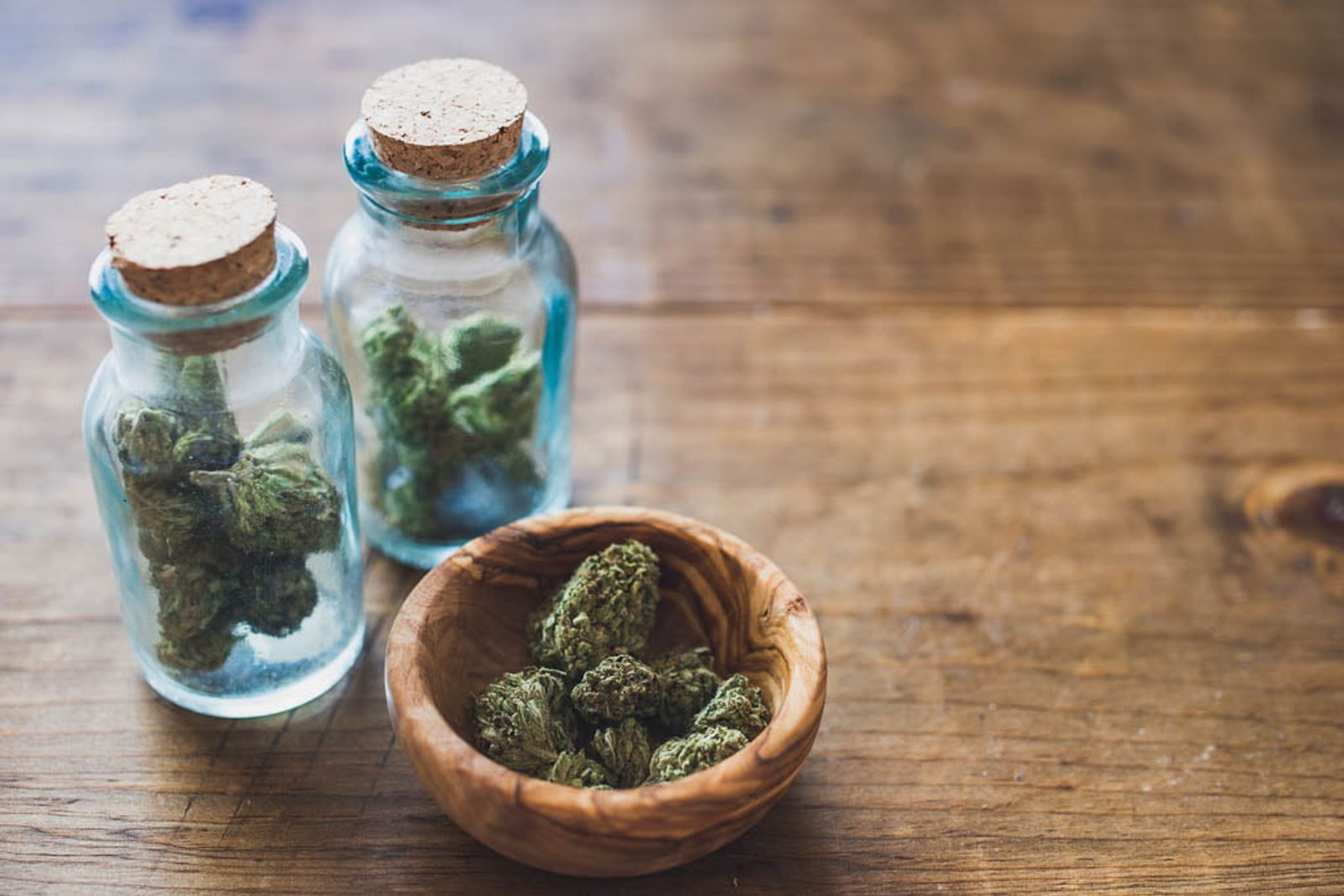 Two glass jars with cork stoppers and wooden bowl containing dried herbs on wooden surface.