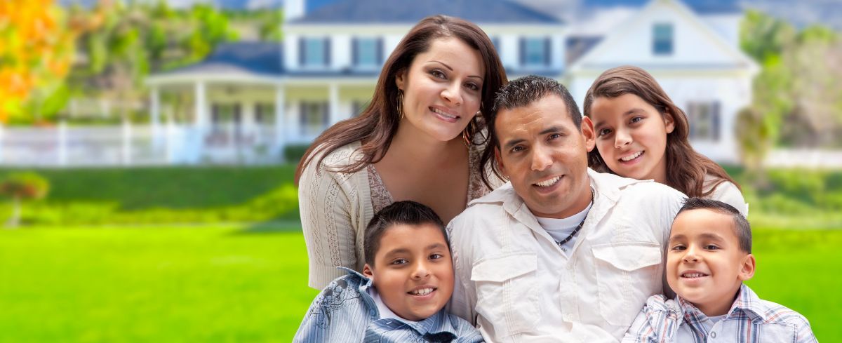 Familia feliz de cinco personas posando junta frente a una gran casa blanca y un césped verde.