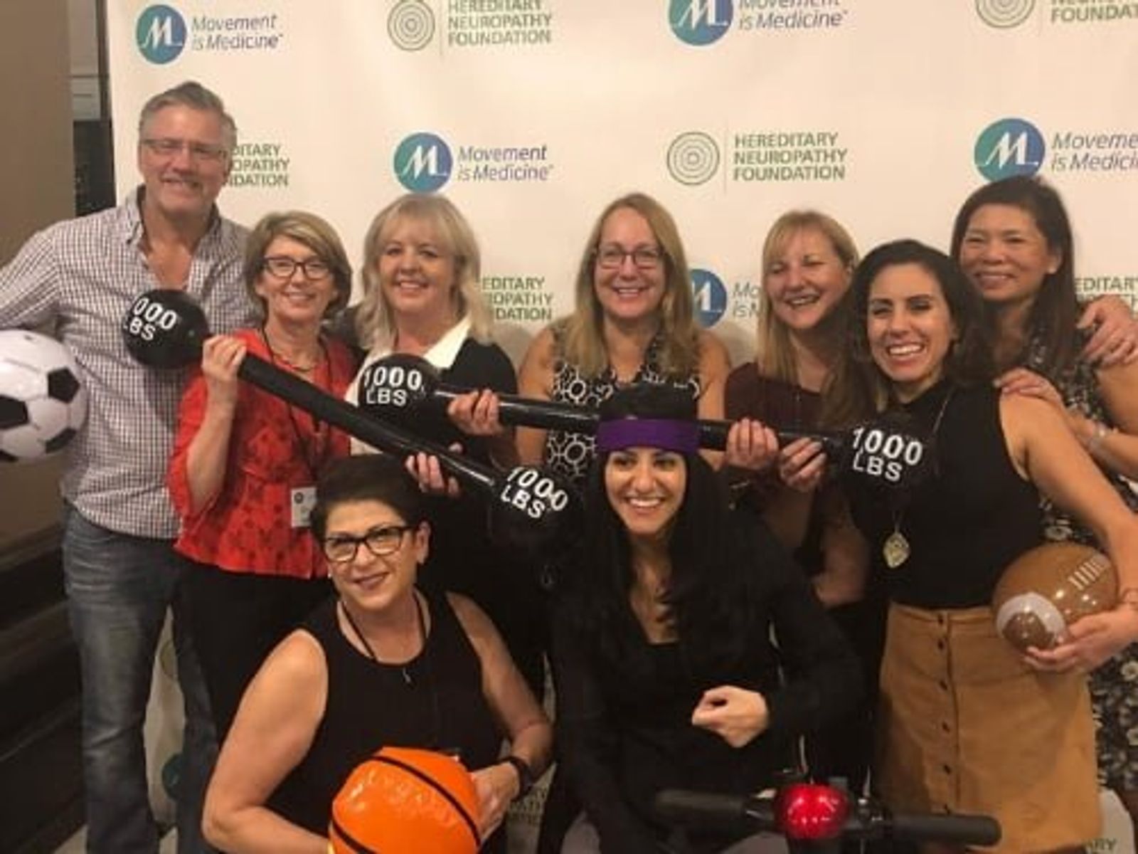 Group of eight people posing with a large rifle prop in front of Movement is Medicine and Hereditary Empathy Foundation backdrops.