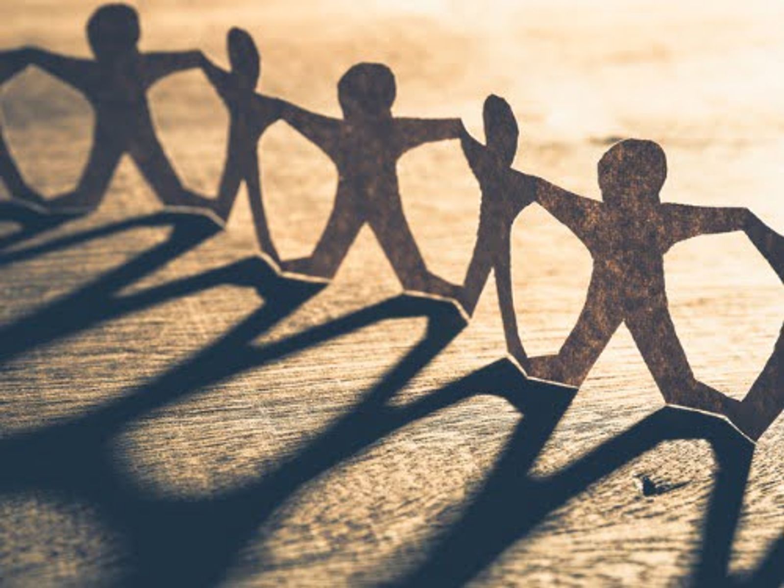 Paper cutout figures holding hands in a chain, casting shadows on wooden surface.