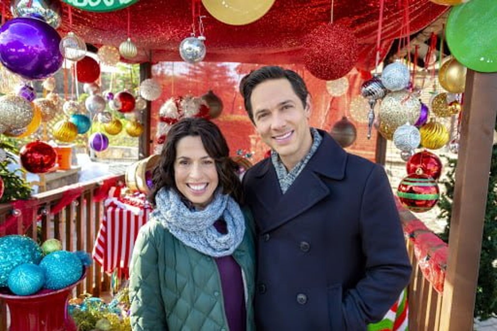 Two people smiling at camera in front of festive holiday decorations with colorful ornaments and garland.