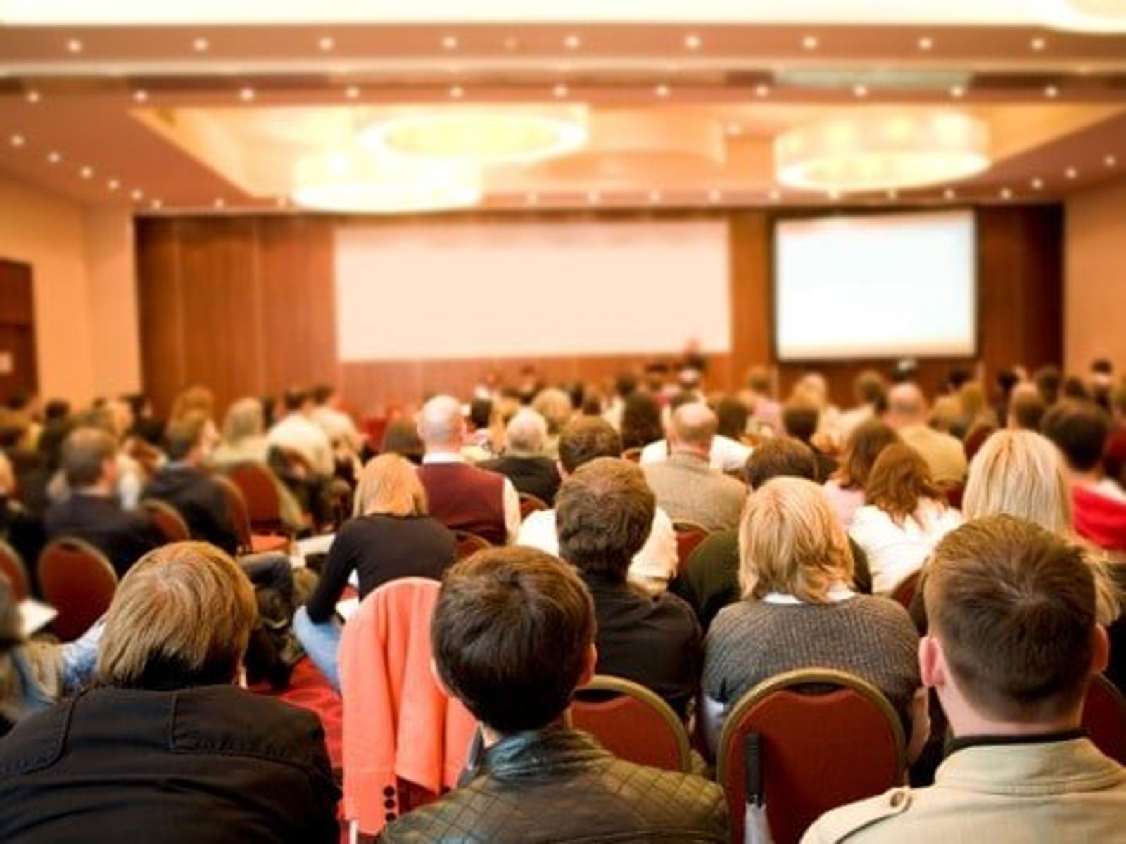 Gran sala de conferencias llena de asistentes sentados frente a pantallas de proyección en la parte delantera.