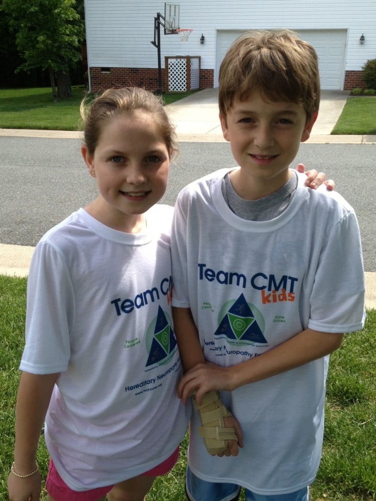 Two children wearing Team CMT Kids t-shirts standing in a driveway in front of a house.