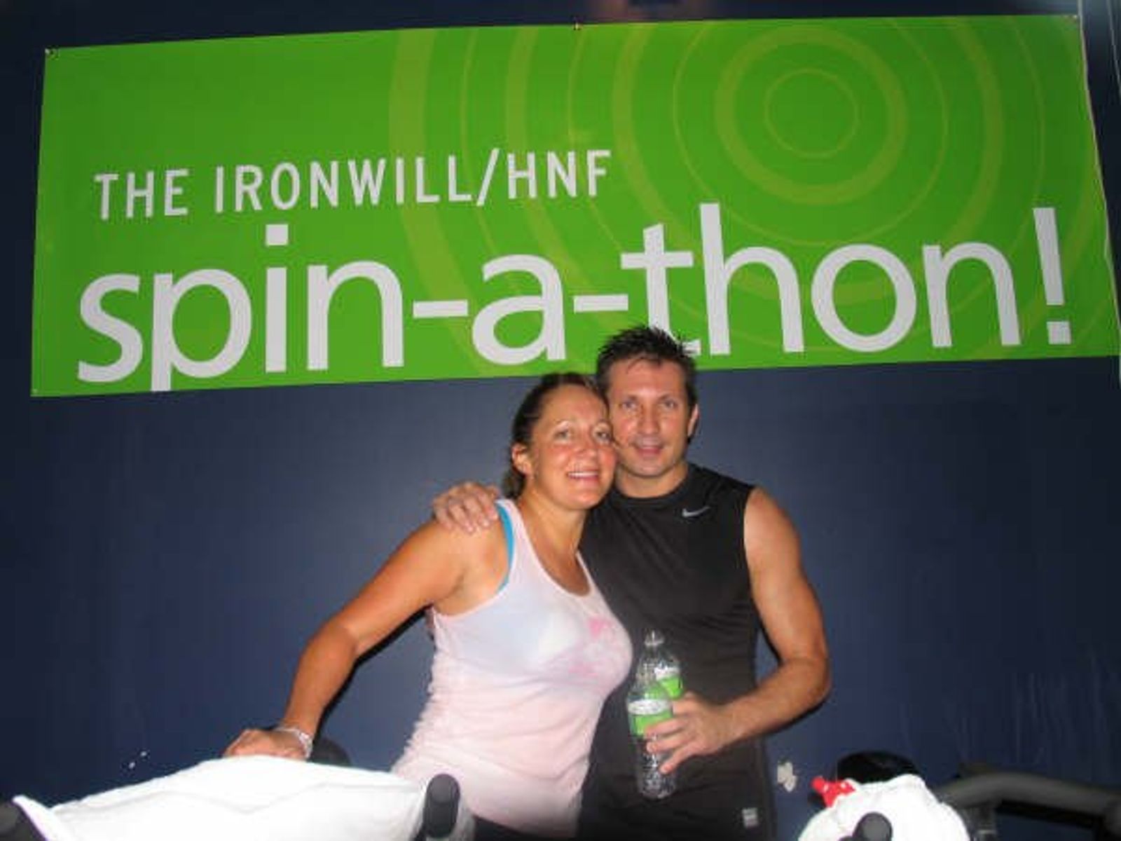 Two people posing together at "THE IRONWILL/HNF spin-a-thon!" event with green banner backdrop.