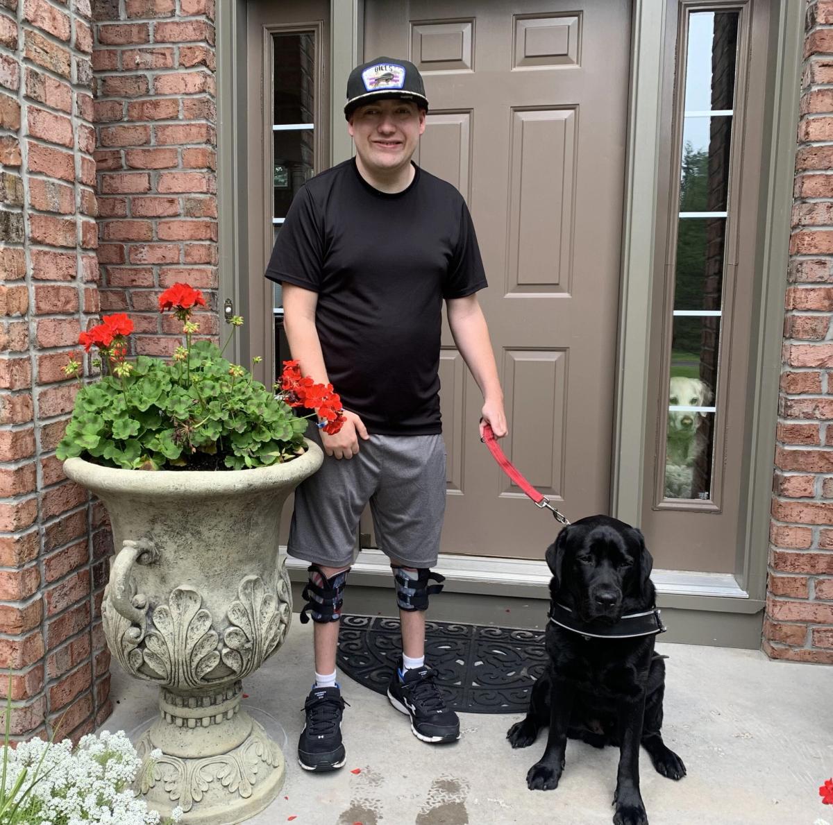 Person in black shirt and cap standing with black dog on leash beside red flowers in front of house.