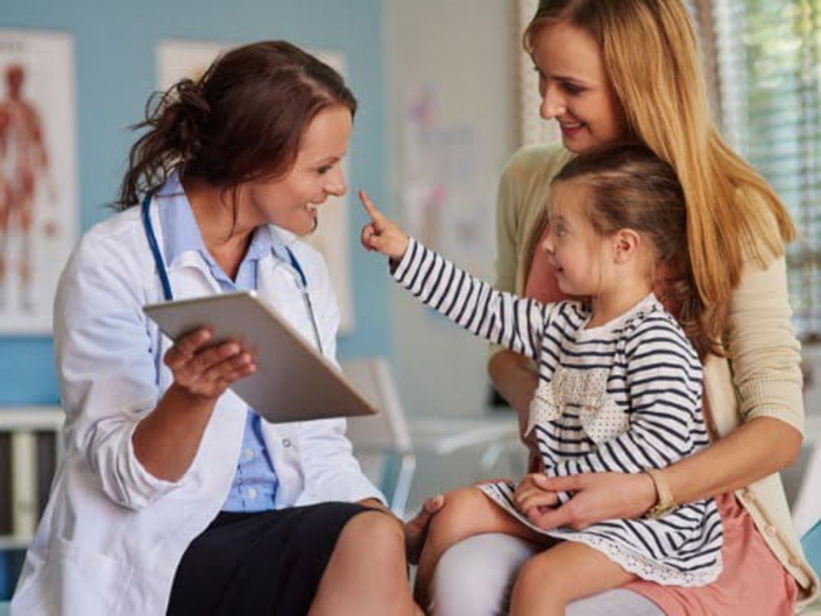 Médica con tableta examina a una niña sonriente mientras su madre está de pie a su lado en un entorno clínico.