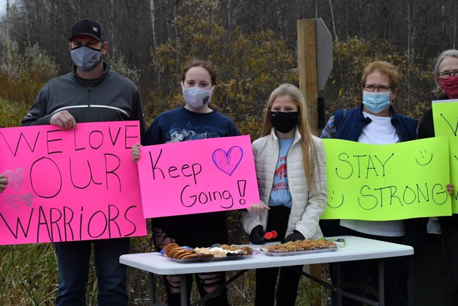 Group of masked people holding signs reading "We love our warriors," "Keep going," and "Stay strong" outdoors.