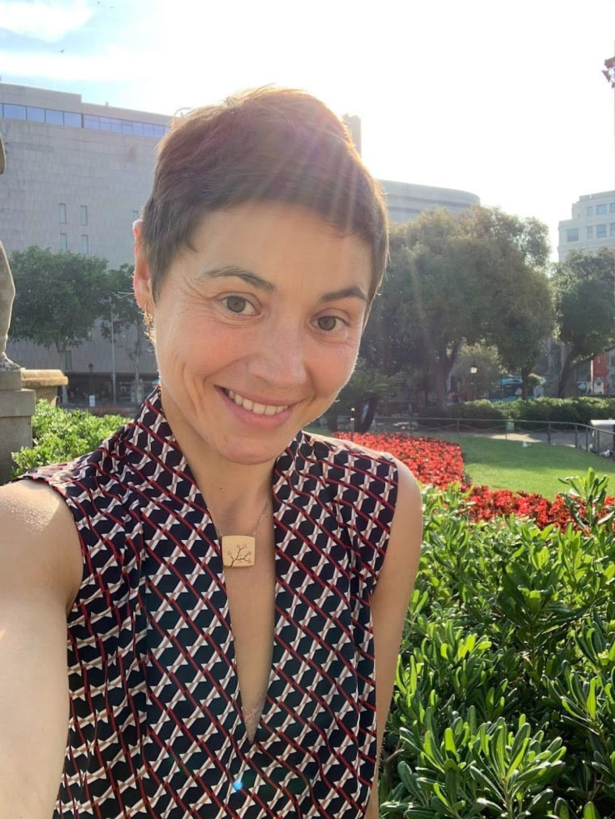 Mujer sonriendo a la cámara en un parque urbano, con flores rojas y edificios detrás de ella.