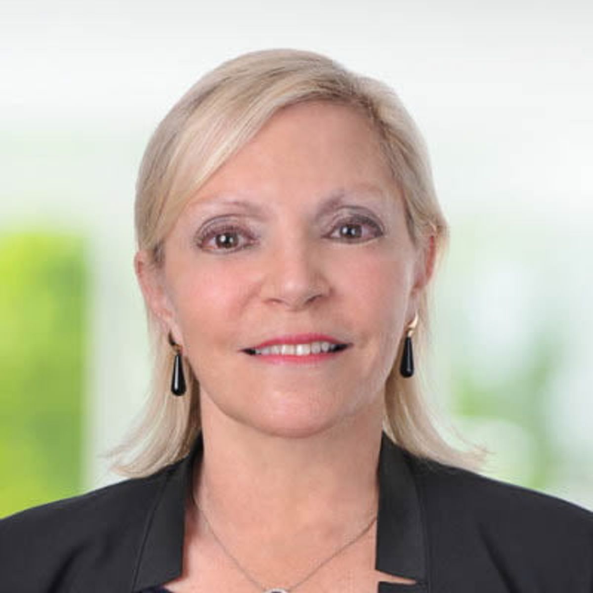 Retrato profesional de una mujer rubia con un blazer negro, frente a un fondo desenfocado.
