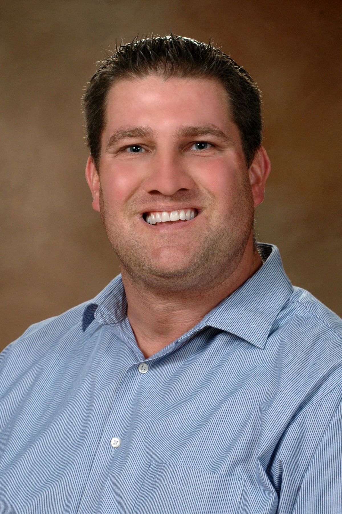 Professional headshot of a man smiling wearing a light blue striped shirt against a brown background.
