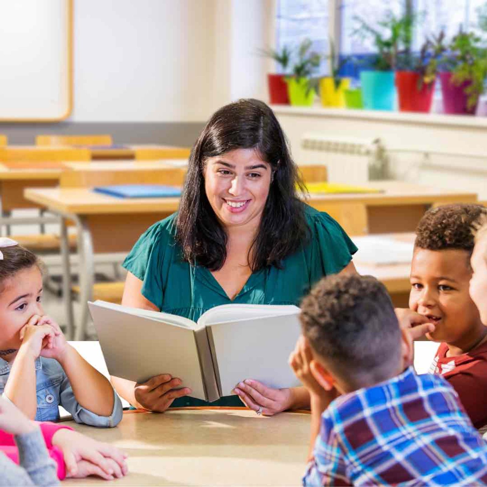 Teacher reading from open book to group of young children seated at wooden table in classroom.