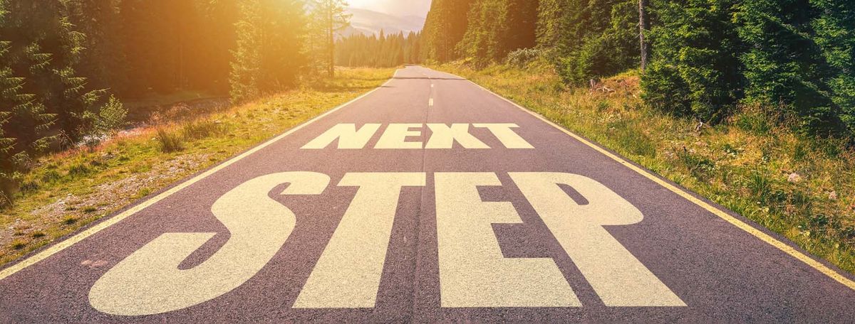 Straight road with "NEXT STEP" painted in white letters, flanked by green trees and sunlight ahead.