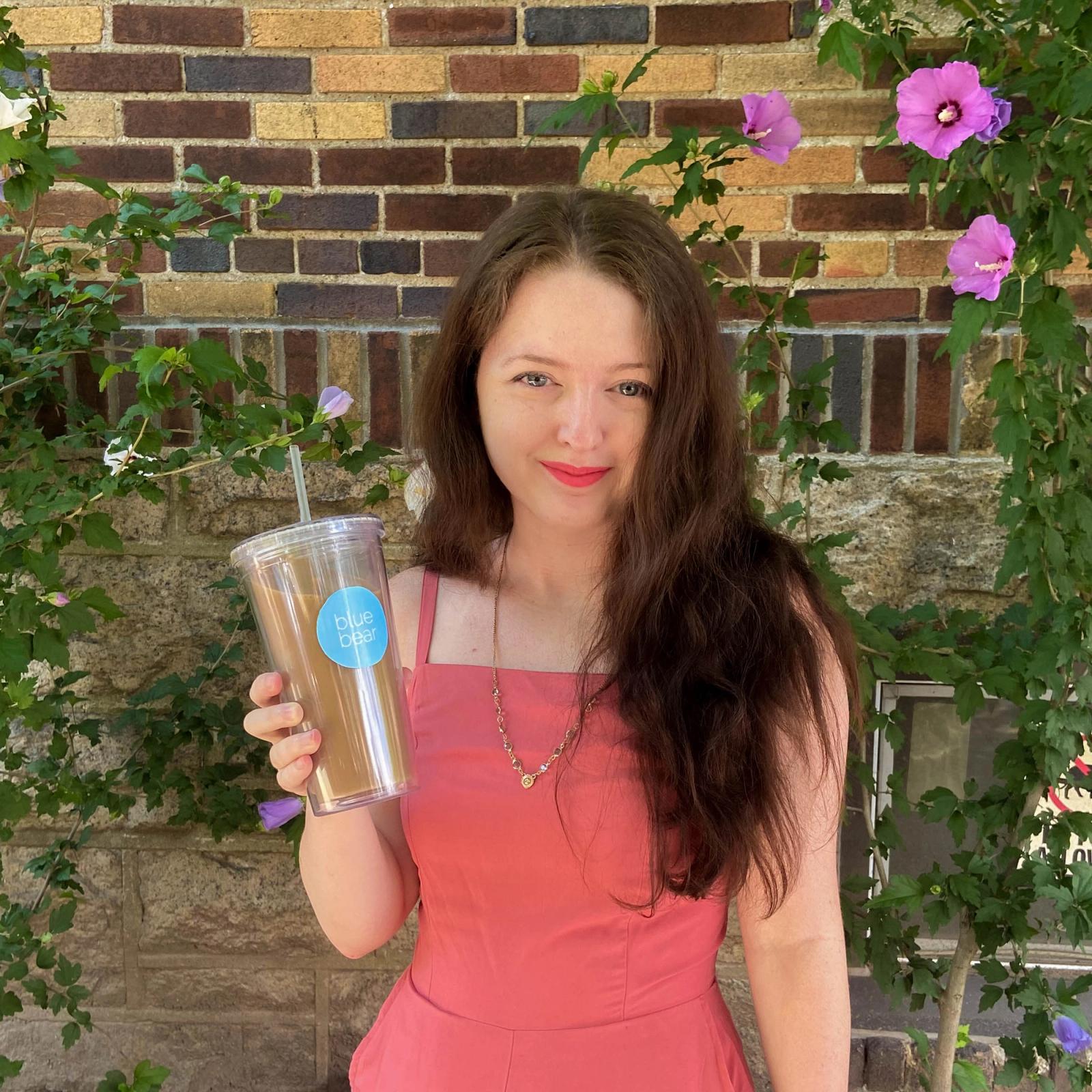 Woman in coral dress holding clear cup with blue snowflake logo against brick wall with pink flowers.