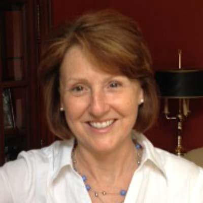 Woman with brown shoulder-length hair smiling at camera, wearing white shirt and blue necklace, burgundy background.