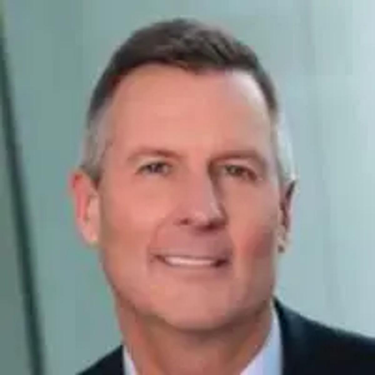 Professional headshot of middle-aged man in dark suit and light blue shirt against light blue background.