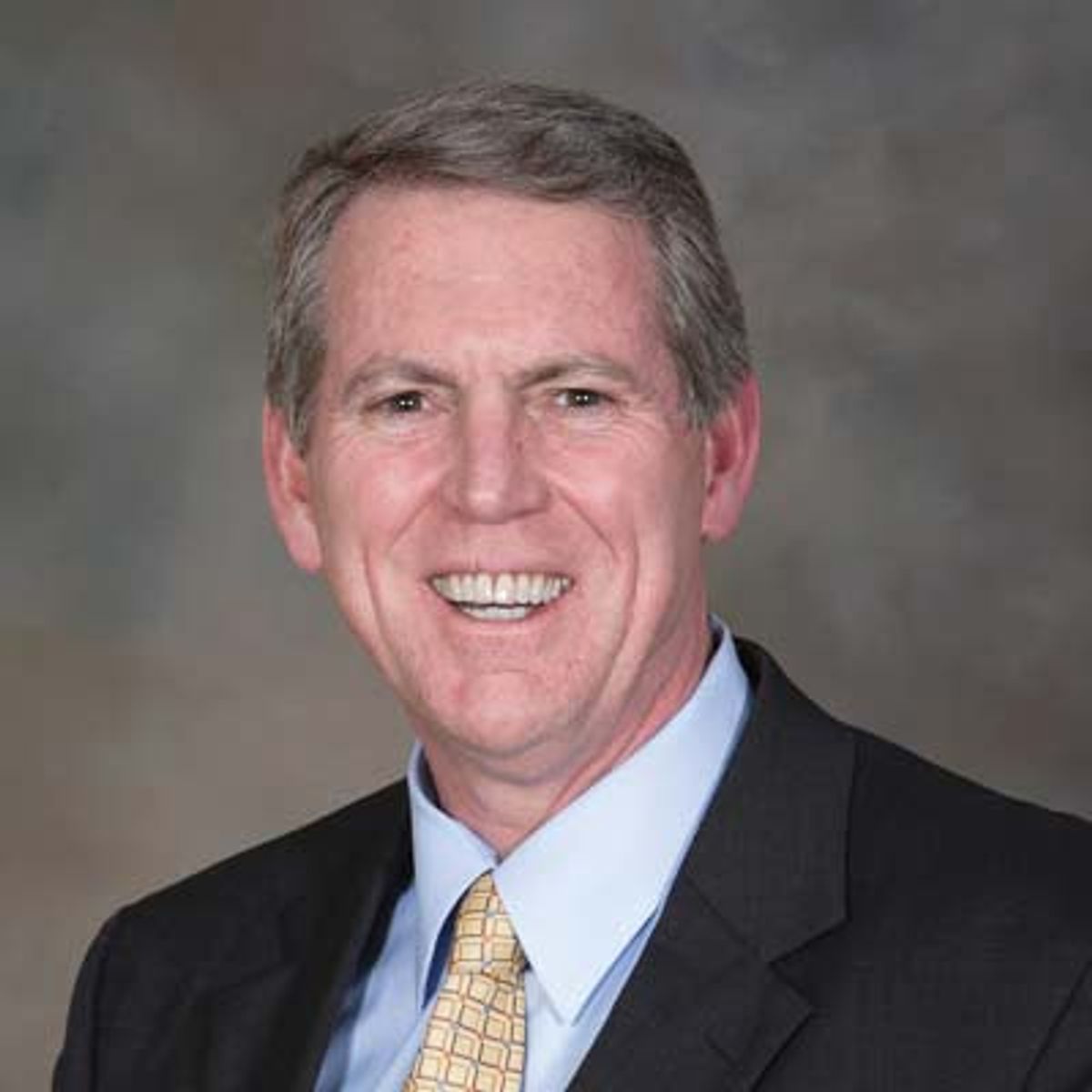 Professional headshot of middle-aged man in dark blazer, light blue shirt, and patterned tie against gray background.