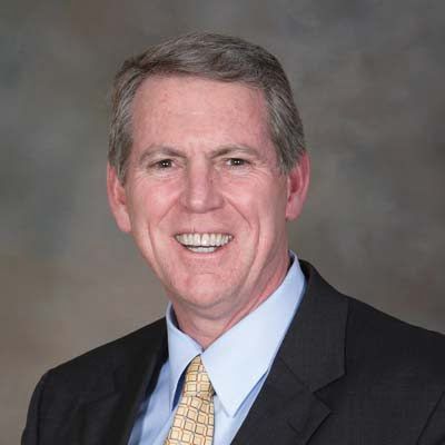 Professional headshot of middle-aged man in dark blazer, light blue shirt, and patterned tie against gray background.