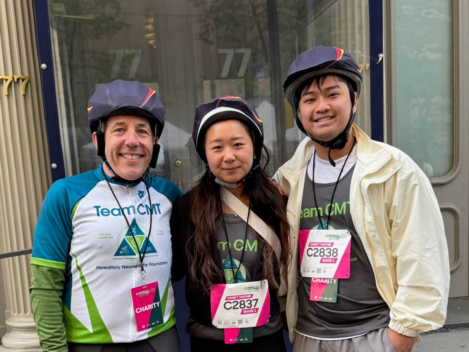 Three people wearing helmets and race bibs stand together in front of a building.
