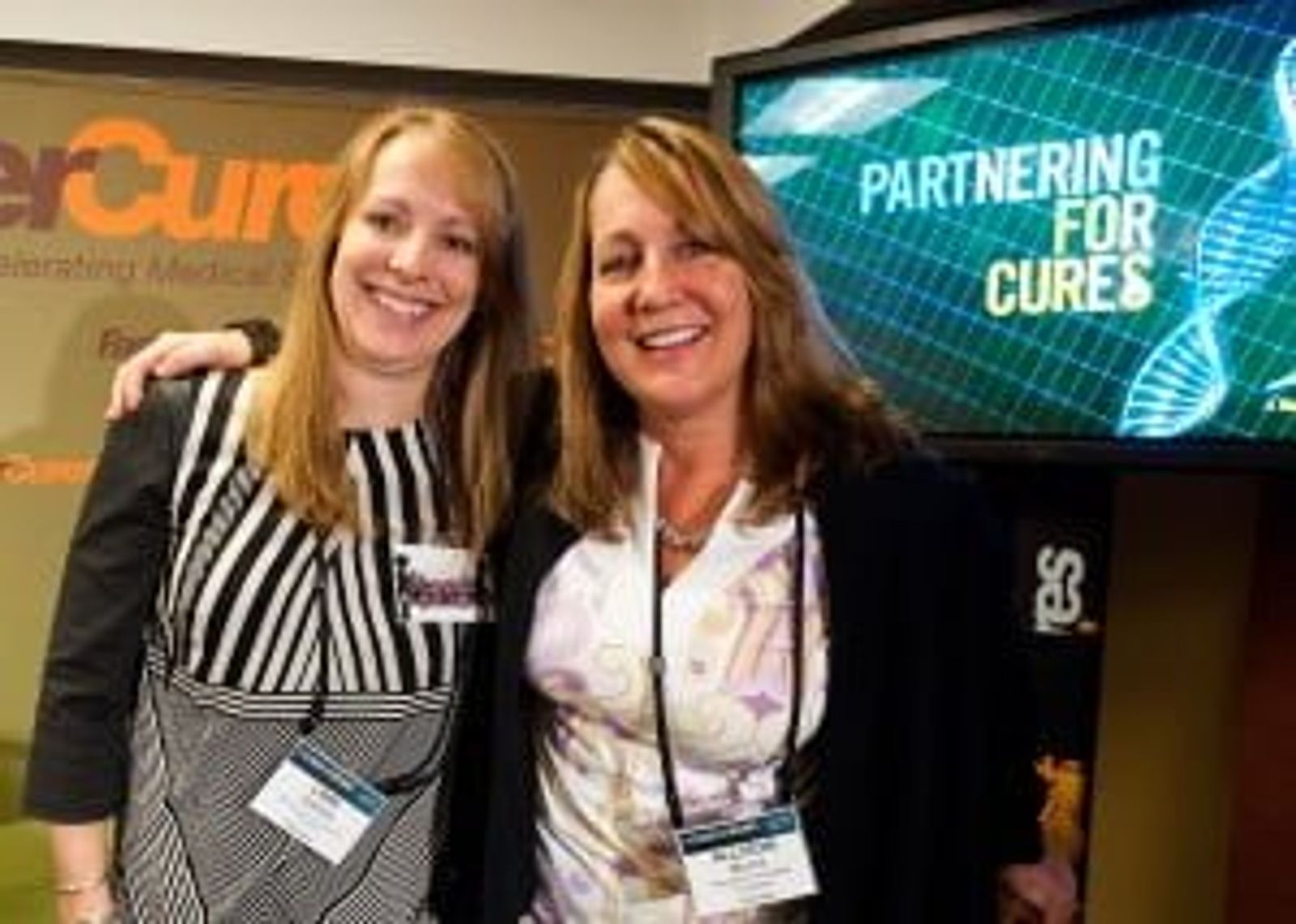 Two women wearing lanyards at conference, "PARTNERING FOR CURES" text visible on screen behind them.