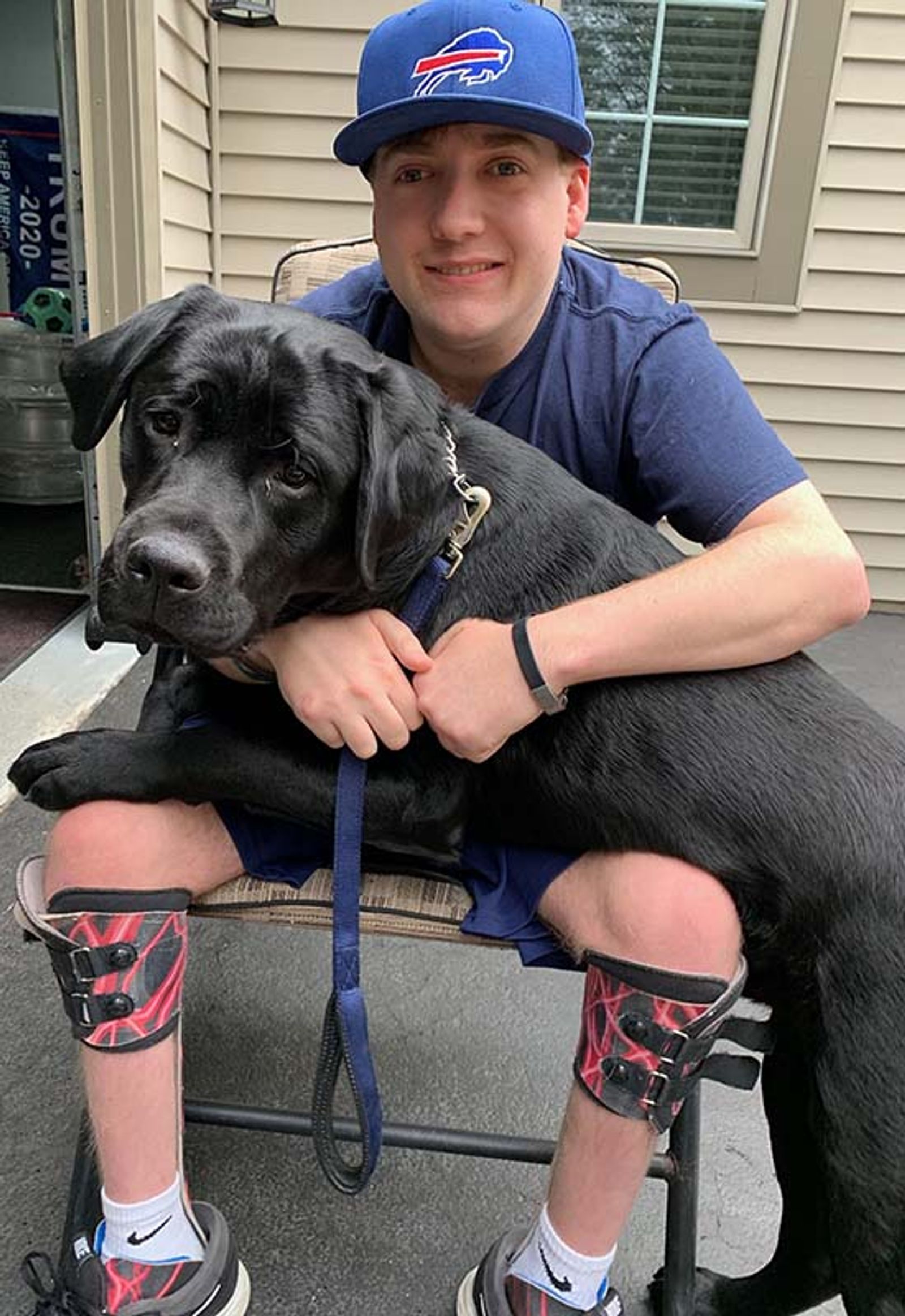 Young man wearing Buffalo Bills cap holding black Labrador on chair outdoors.