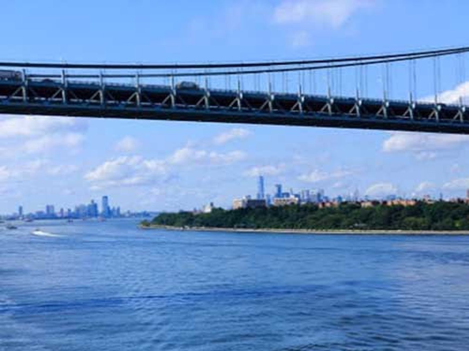 Suspension bridge spanning river with Manhattan skyline visible in background under blue sky.