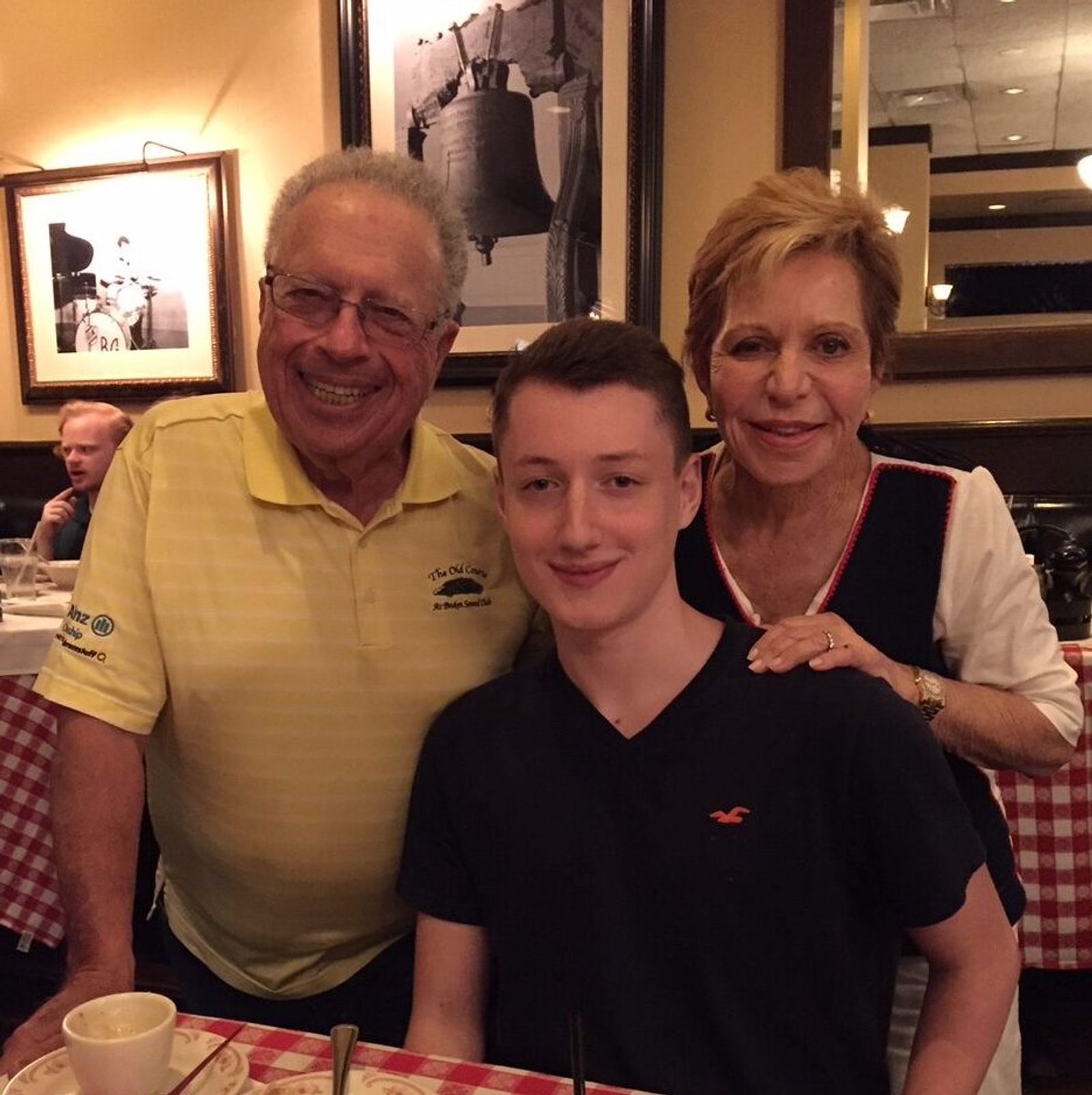 Three people posing together at a restaurant table with checkered tablecloth and framed photos on wall behind them.
