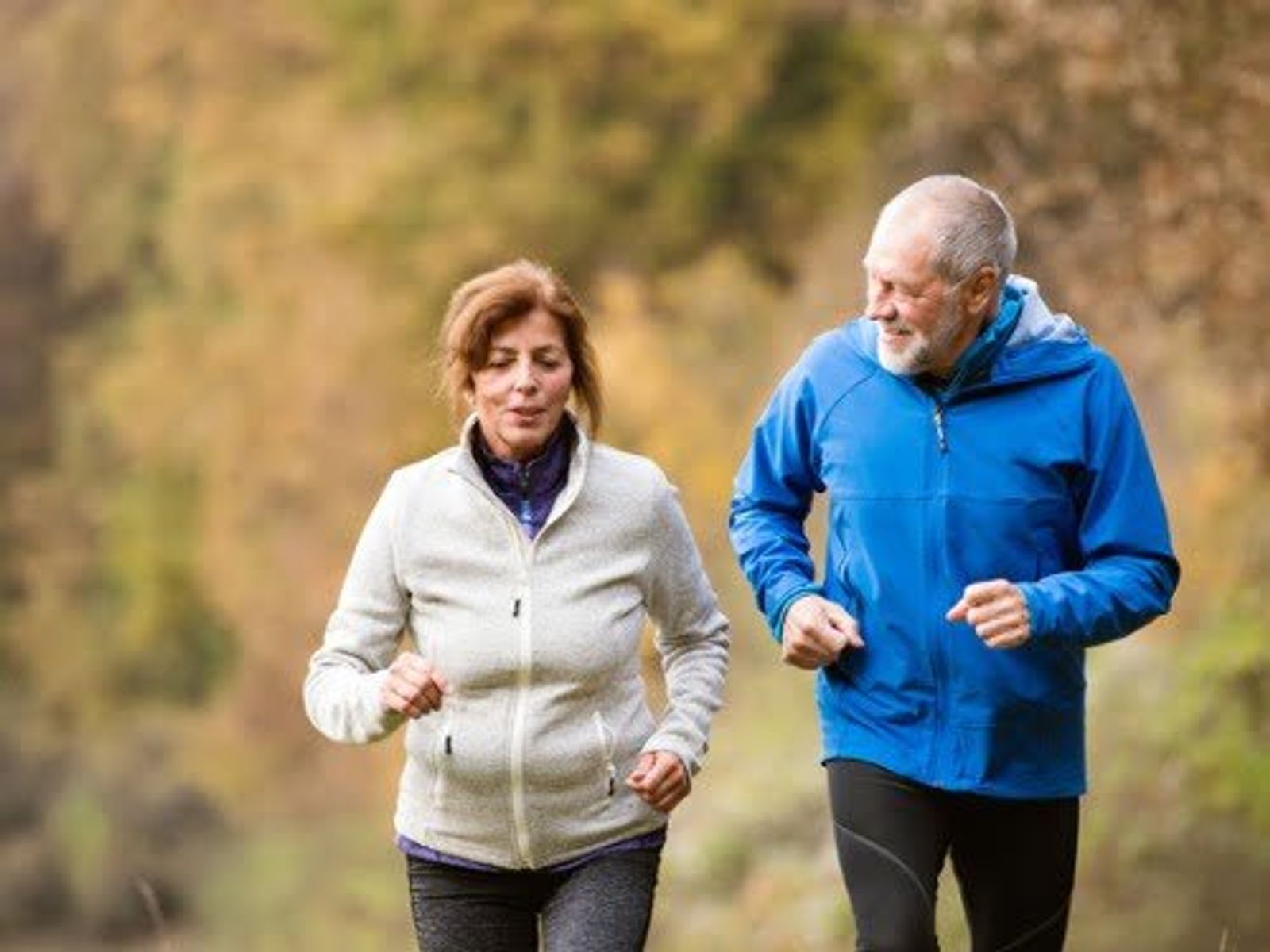 Two older adults jogging outdoors on a path with autumn foliage in background.