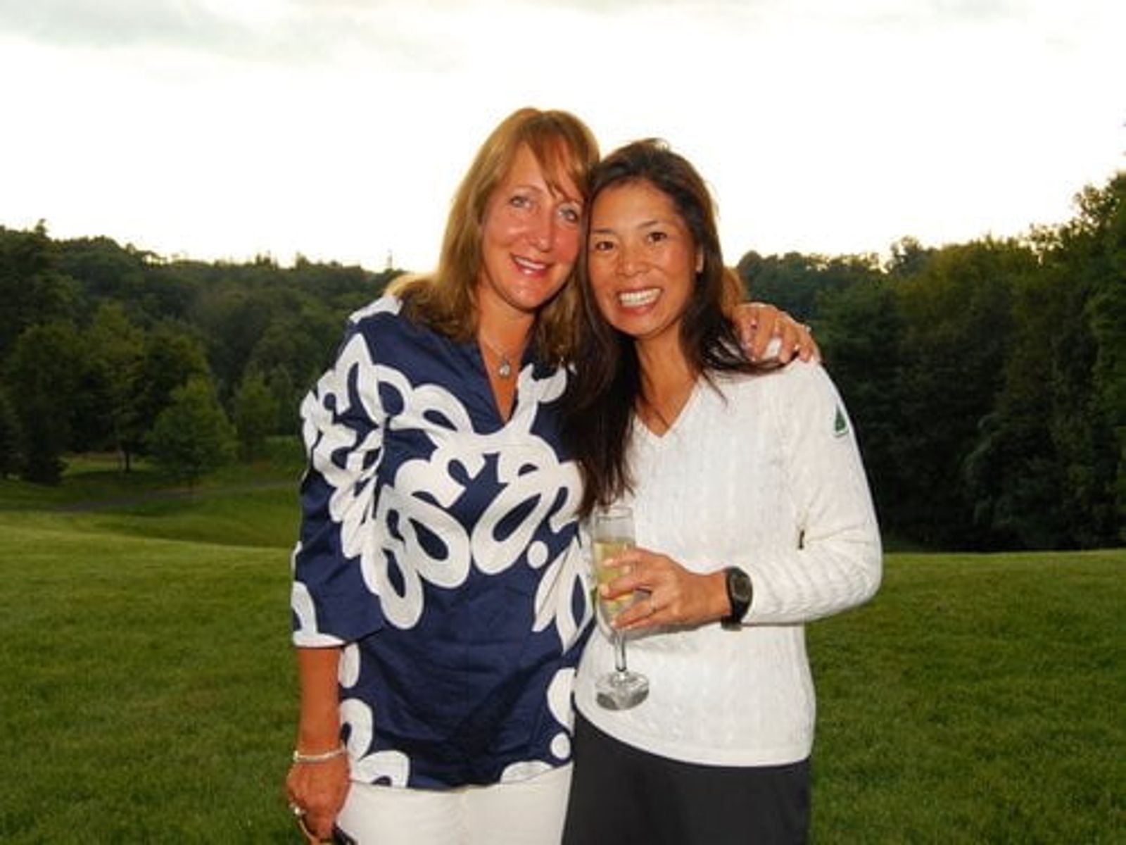 Two women smiling outdoors in a grassy field with trees in background.