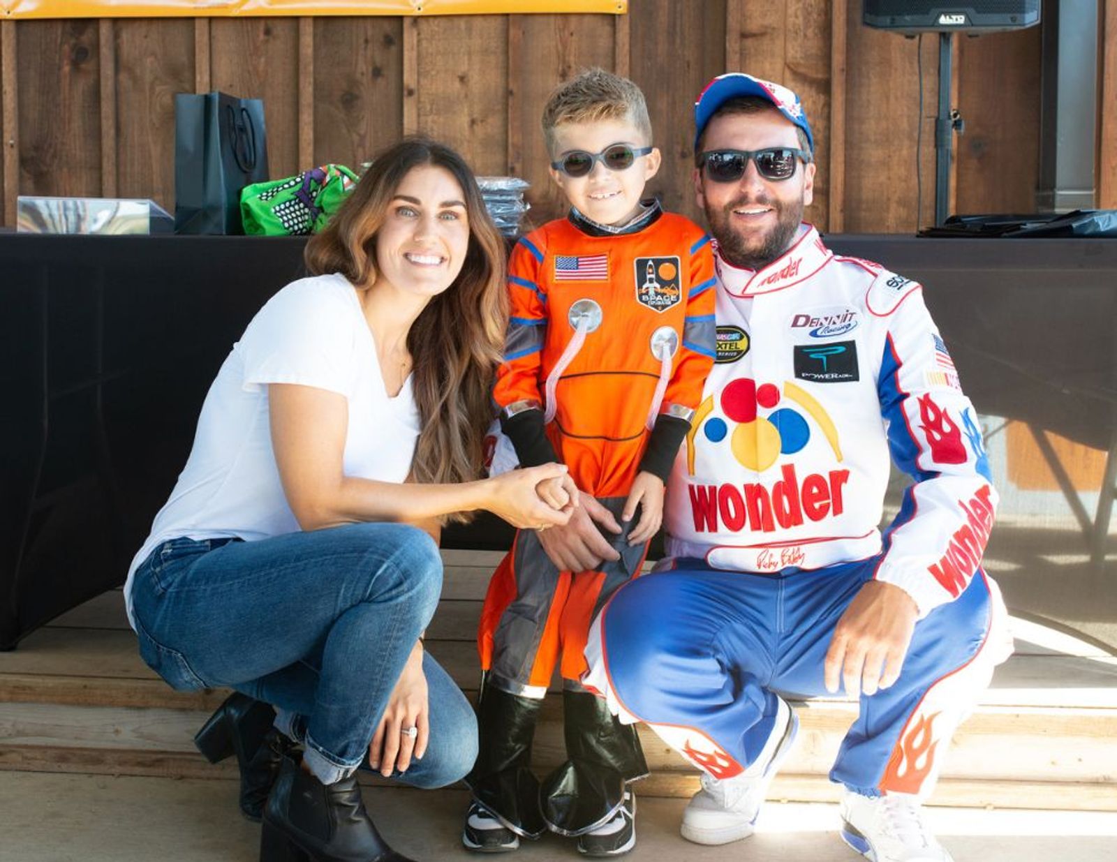 Three people posing indoors: woman in white shirt and jeans, child in orange astronaut suit, man in white and blue racing suit with "Wonder" logo.