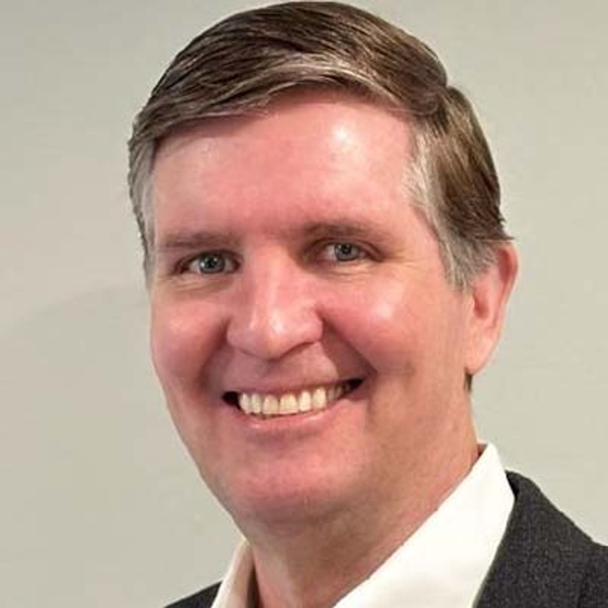 Professional headshot of a middle-aged man smiling at camera, wearing dark jacket and white shirt.
