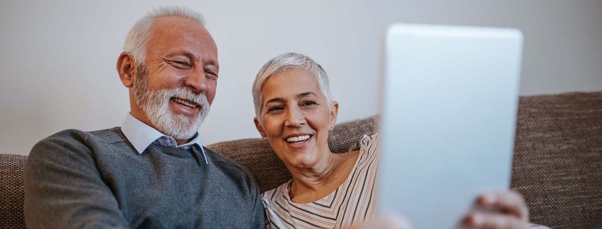 Senior couple smiling while holding tablet together on couch.