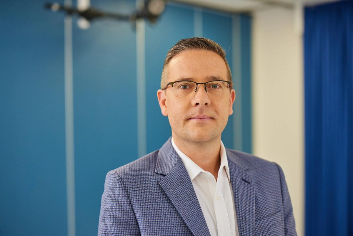 Professional man wearing glasses and blue blazer against blue and white striped background.