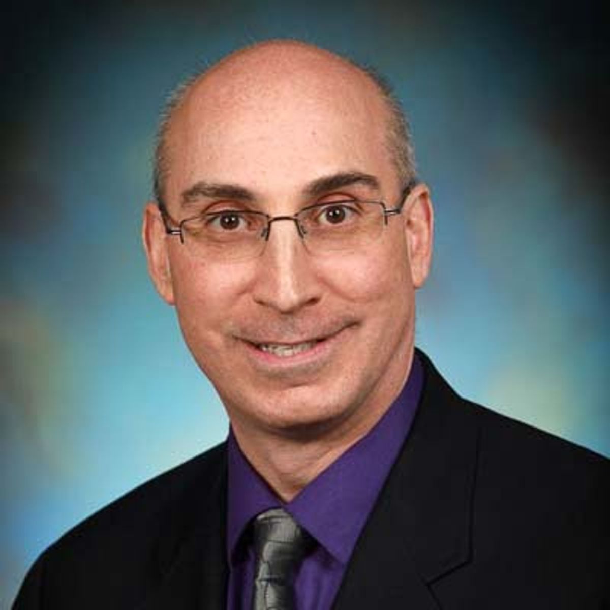 Professional headshot of a bald man wearing glasses, purple shirt, and dark blazer against blue background.