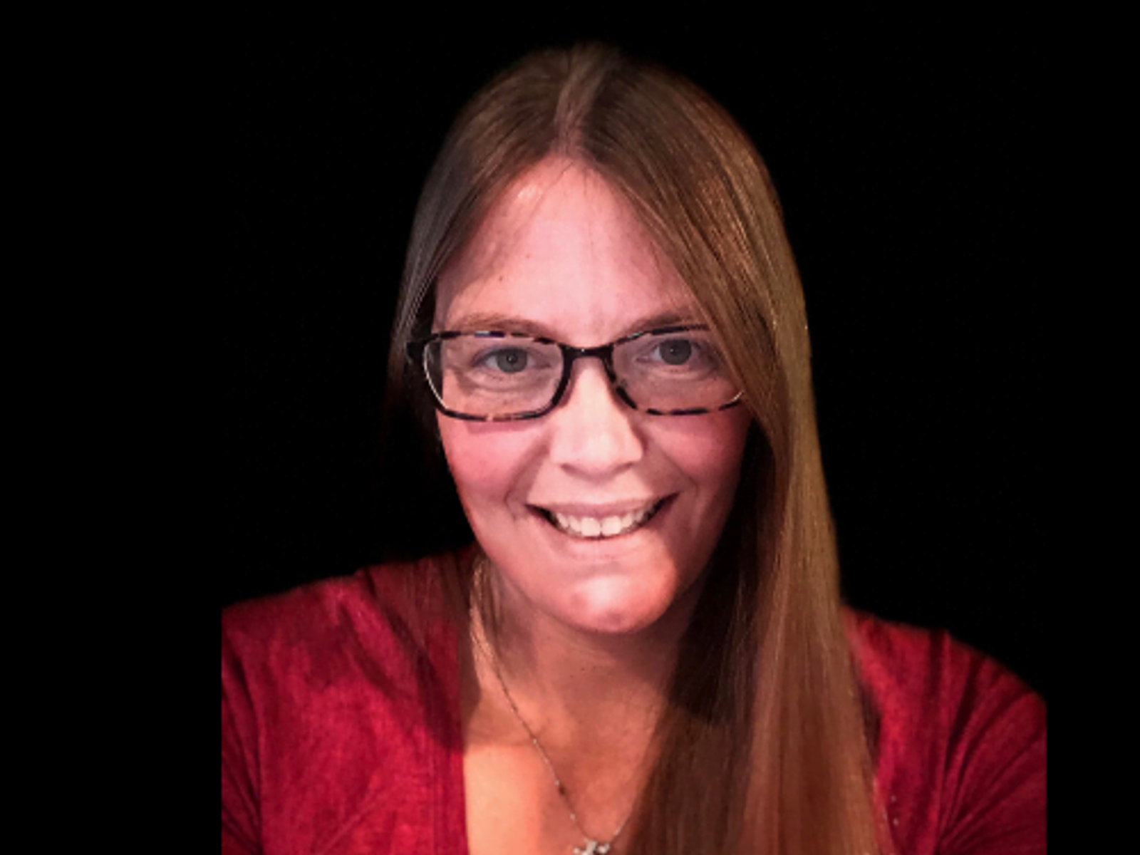 Woman with long reddish-brown hair and glasses smiling against black background, wearing red top.