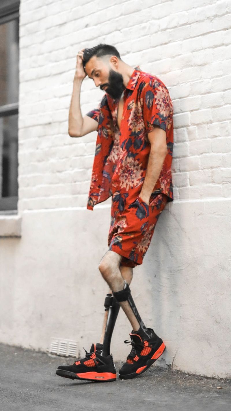 Hombre con camisa y pantalones cortos rojos con estampado floral, apoyado contra una pared de ladrillo blanco, con zapatillas negras y naranjas.