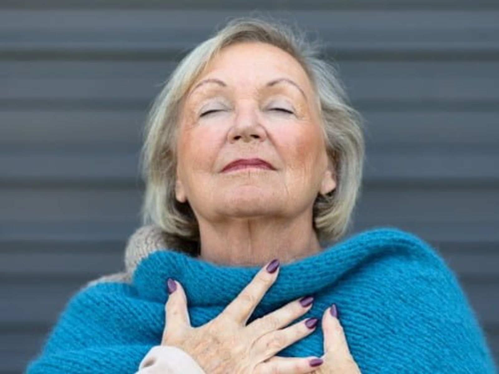 Older woman with gray hair wearing blue sweater, eyes closed, hand on chest against dark background.