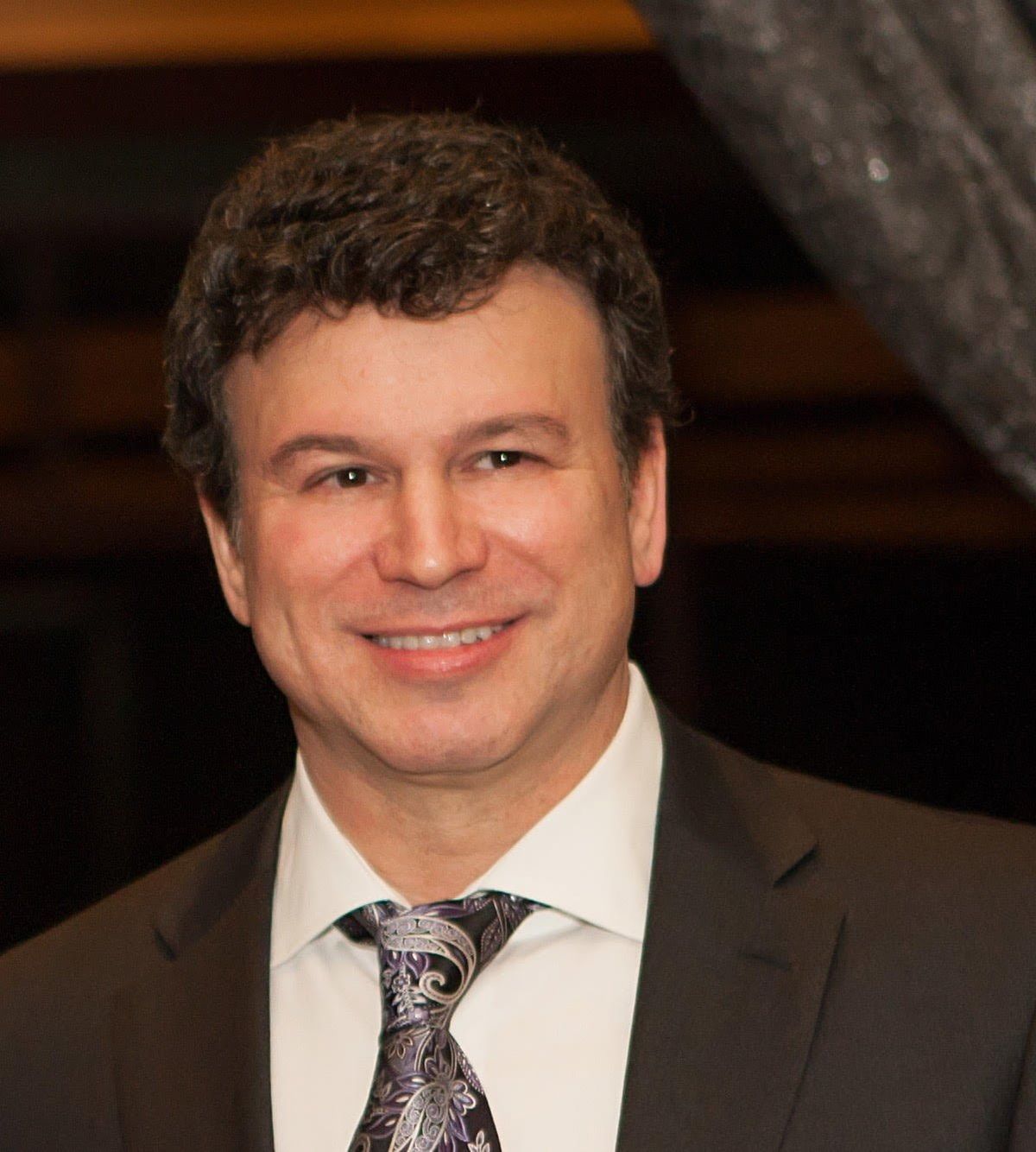 Professional headshot of man in dark suit, white shirt, and patterned tie, smiling at camera.