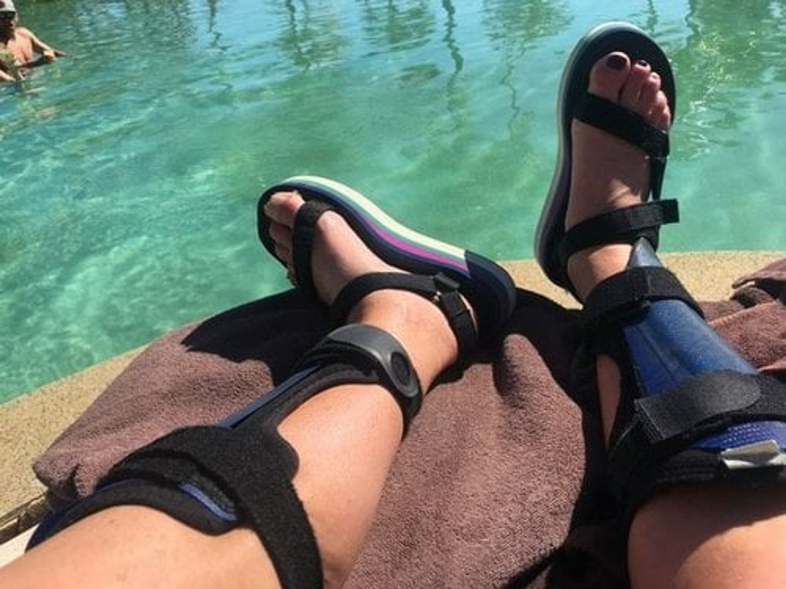 Person wearing black sandals sitting by turquoise water with towel on lap.