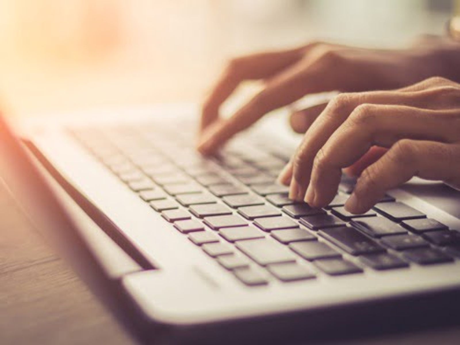 Hands typing on a white laptop keyboard with warm sunlight in background.