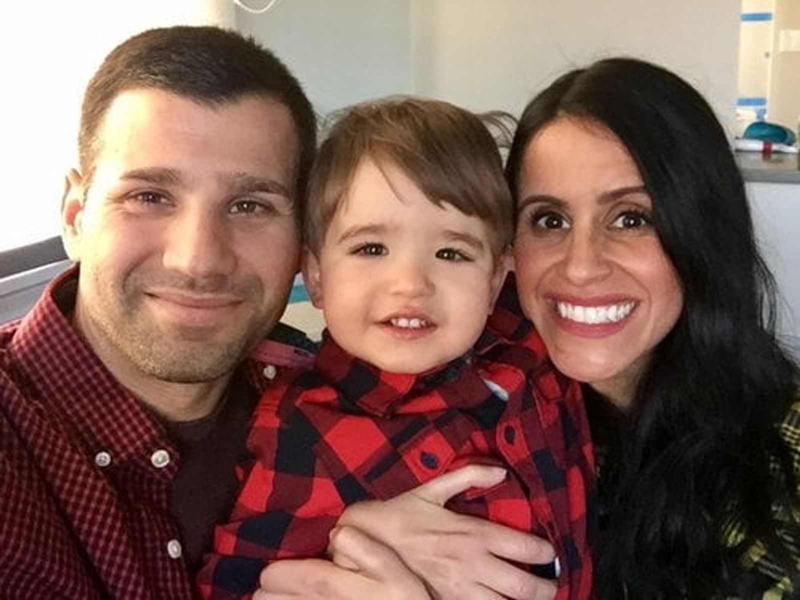 Three people smiling at camera: man in red plaid shirt, young child in red plaid shirt, woman with long dark hair.