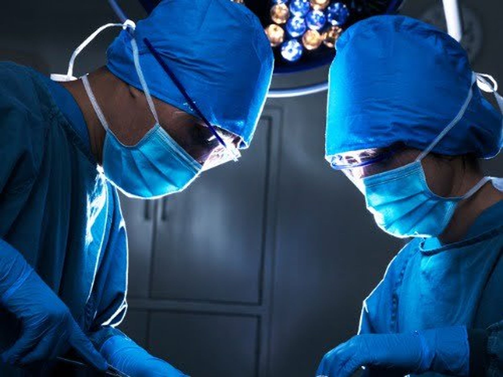 Two surgeons in blue surgical attire performing operation under overhead surgical lights.
