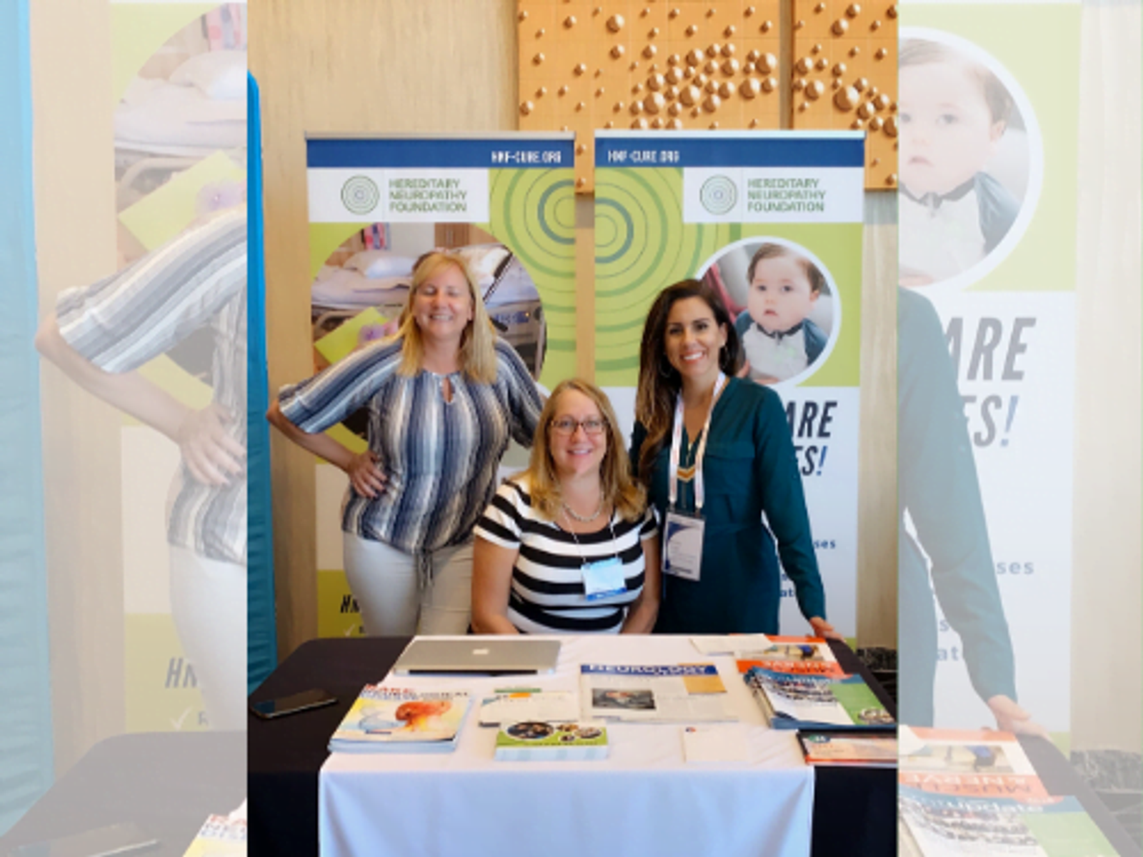 Three women at exhibition booth with informational materials and branded backdrop signage.