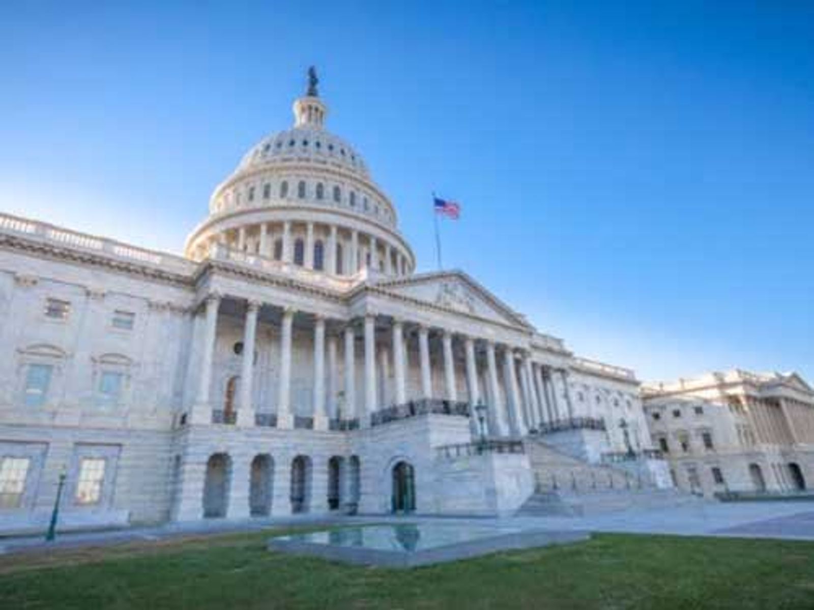 United States Capitol Building with dome, columns, and American flag on clear day.
