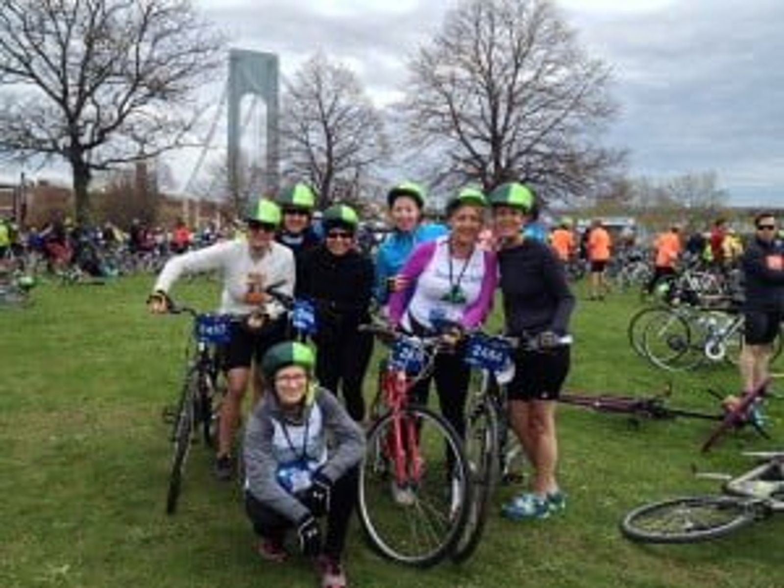Group of cyclists wearing neon helmets posing in park with Verrazano Bridge visible in background.