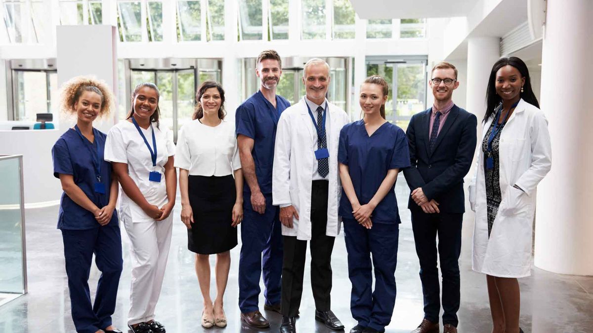 Healthcare team of eight professionals in medical uniforms and coats standing together in modern hospital corridor.