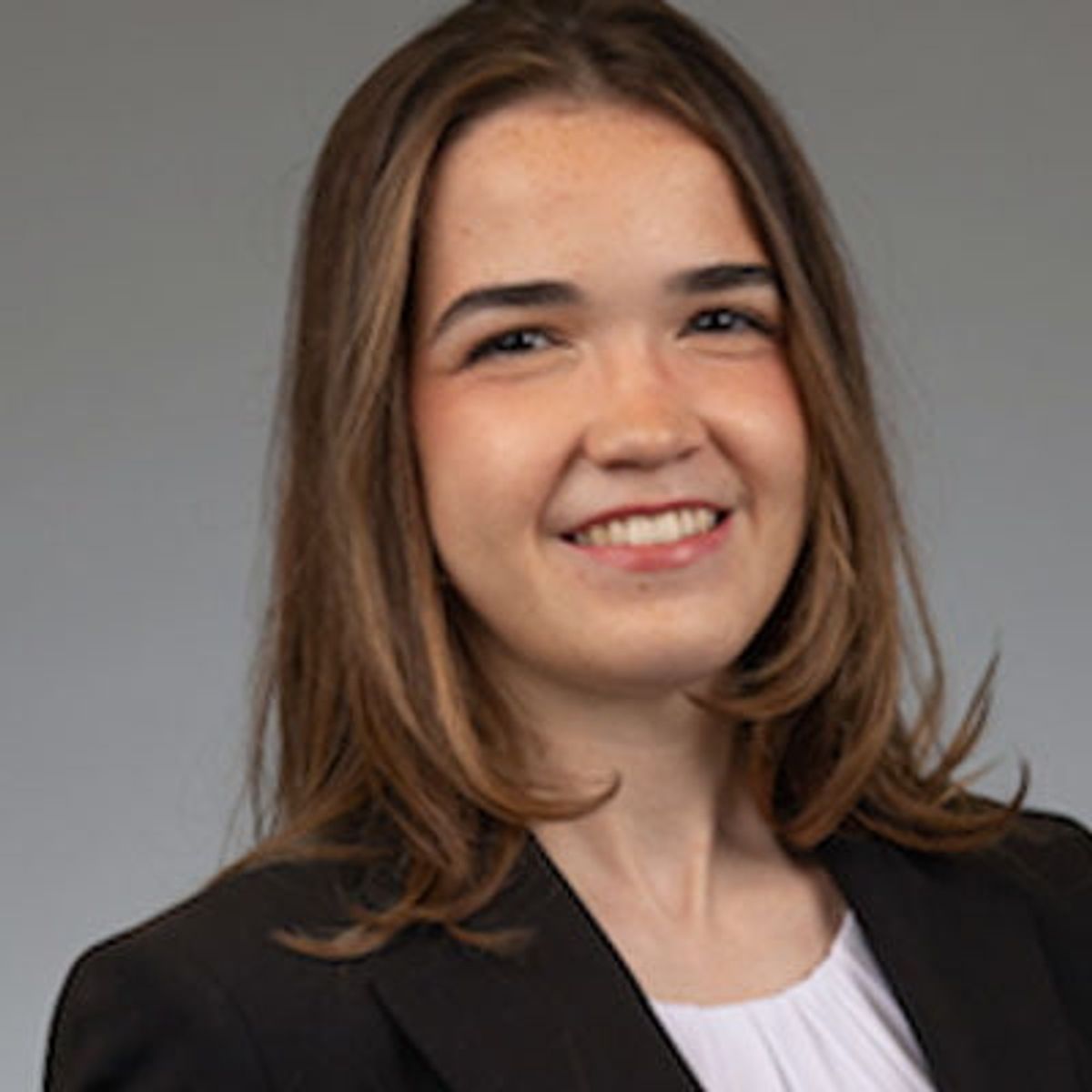 Retrato profesional de una mujer con cabello castaño que lleva un blazer negro y una prenda blanca, sonríe sobre un fondo gris.