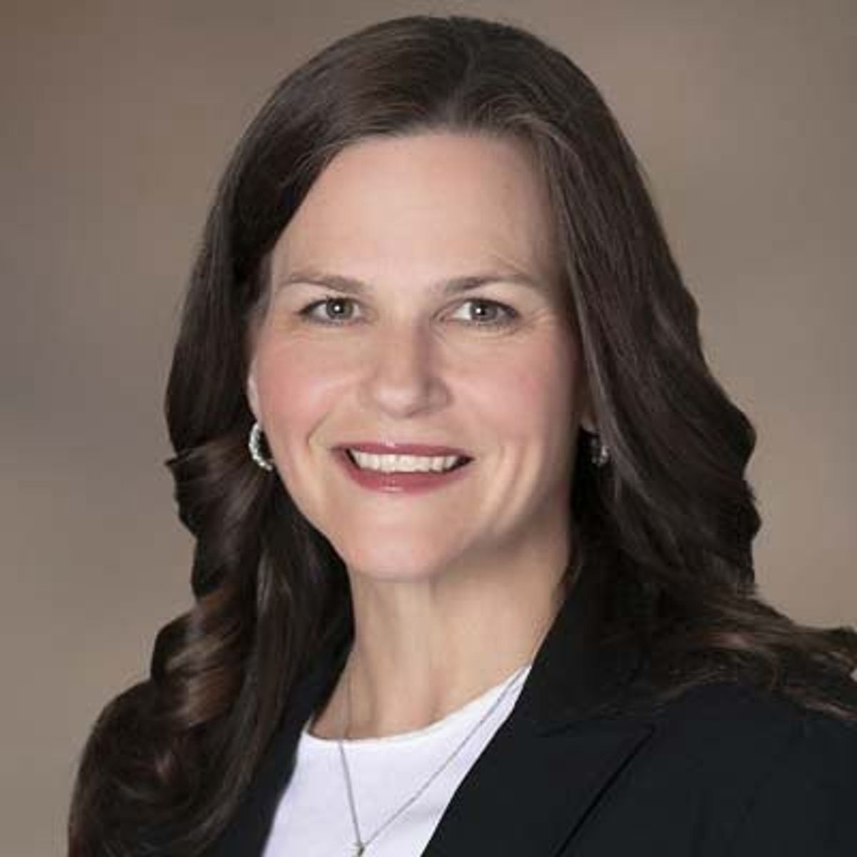 Professional headshot of a woman with long dark hair wearing a black blazer and white top against a neutral background.