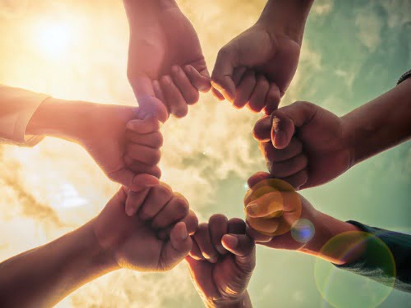 Multiple hands joined together in circle against bright sky and sunlight.