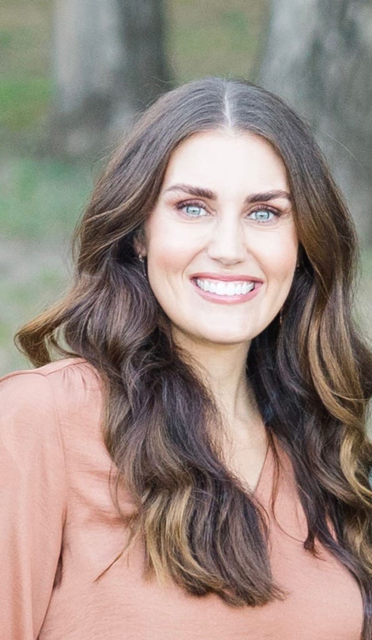 Woman with long dark wavy hair and blue eyes, wearing a peach-colored top, smiling at camera.